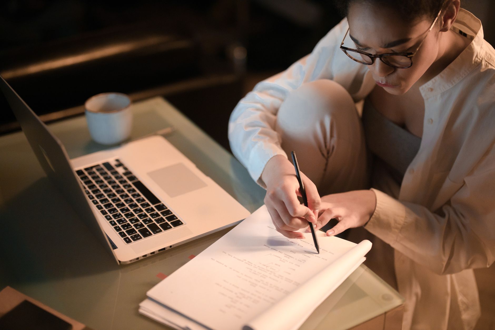A student in glasses marks up printed notes beside an open laptop and a coffee cup late in the evening