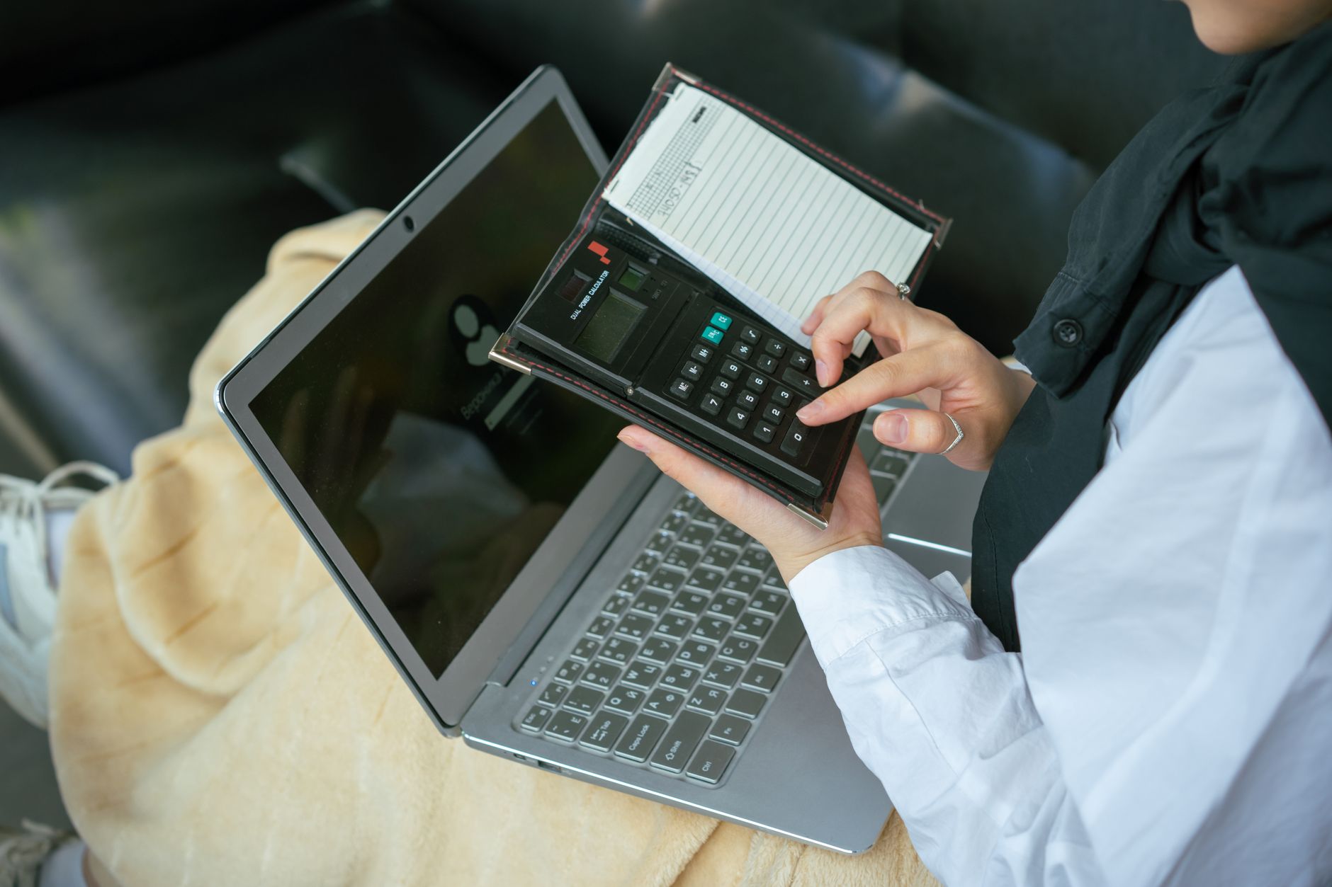 A student in a white shirt taps figures into a pocket calculator held above an open laptop