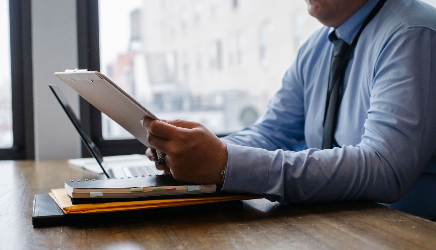 A manager in a shirt and tie reads a report on a clipboard at a desk beside a laptop and a stack of folders