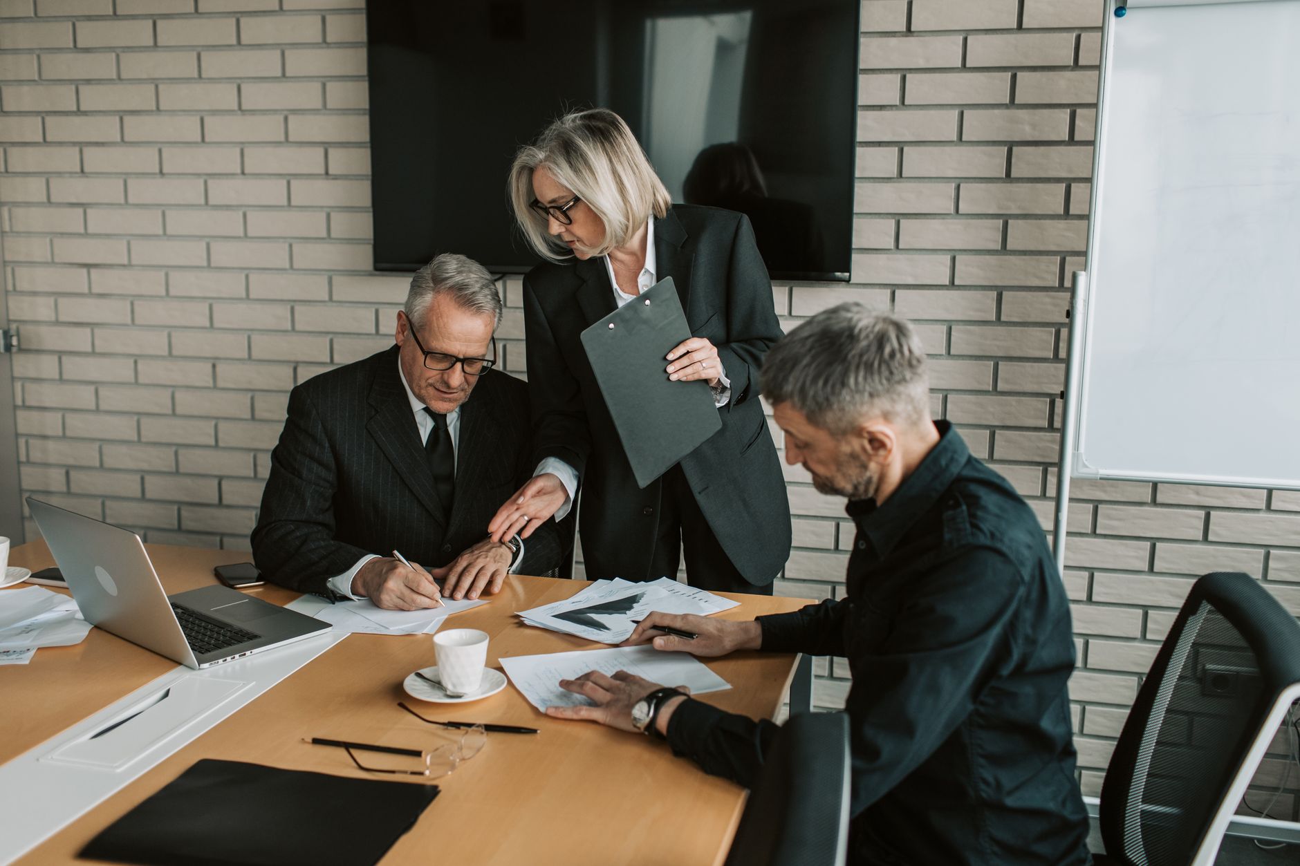 Three senior managers review and sign documents at a conference table while a colleague holding a clipboard points to a page