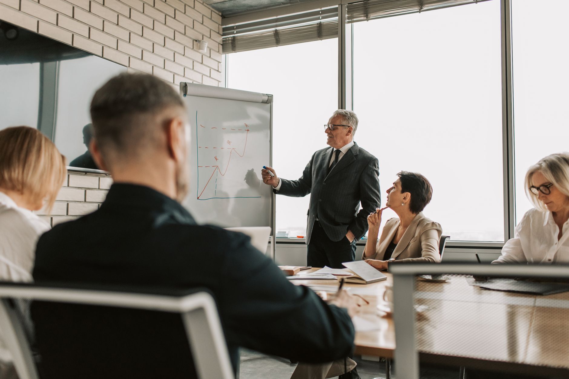 A senior executive sketches a rising chart on a flip chart for colleagues around a conference table