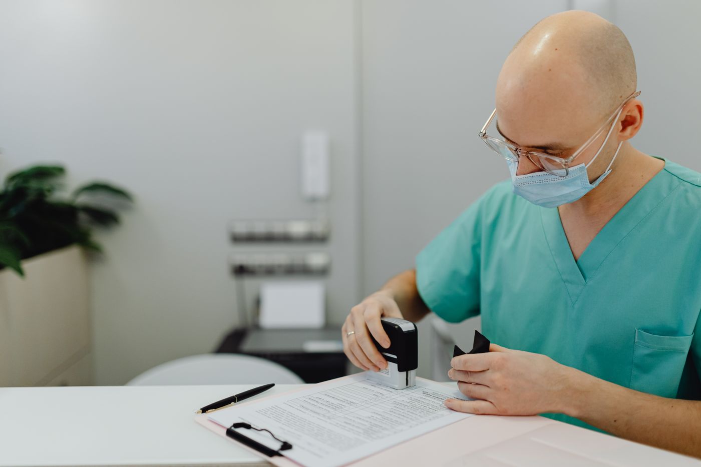 A health worker in scrubs and a face mask stamps a form on a clipboard at a desk