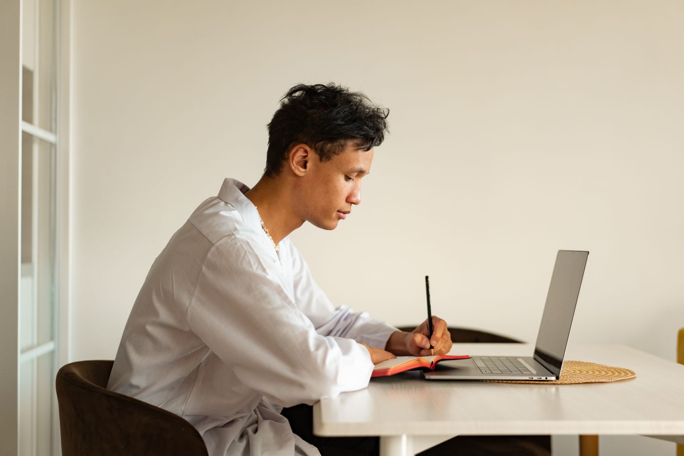 A student in a white shirt writes notes beside an open laptop at a bare table