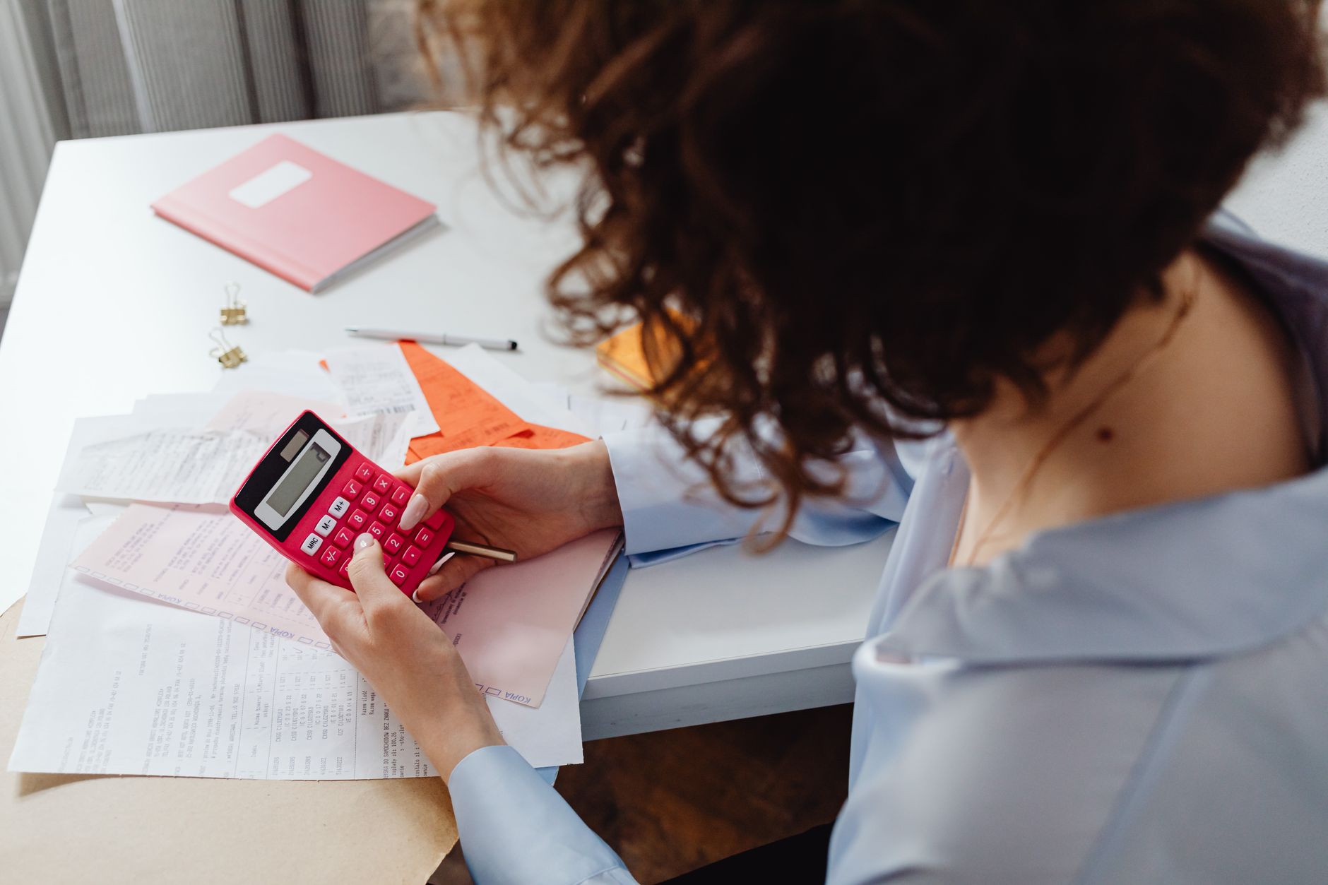 A woman works through bills and receipts with a pink calculator at a white desk