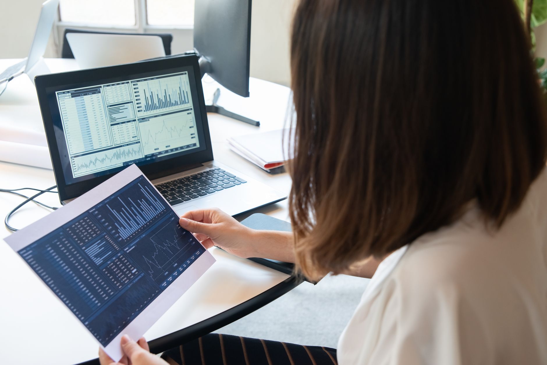 A manager holds a printed report of bar and line charts while the same dashboard is open on a laptop at her desk