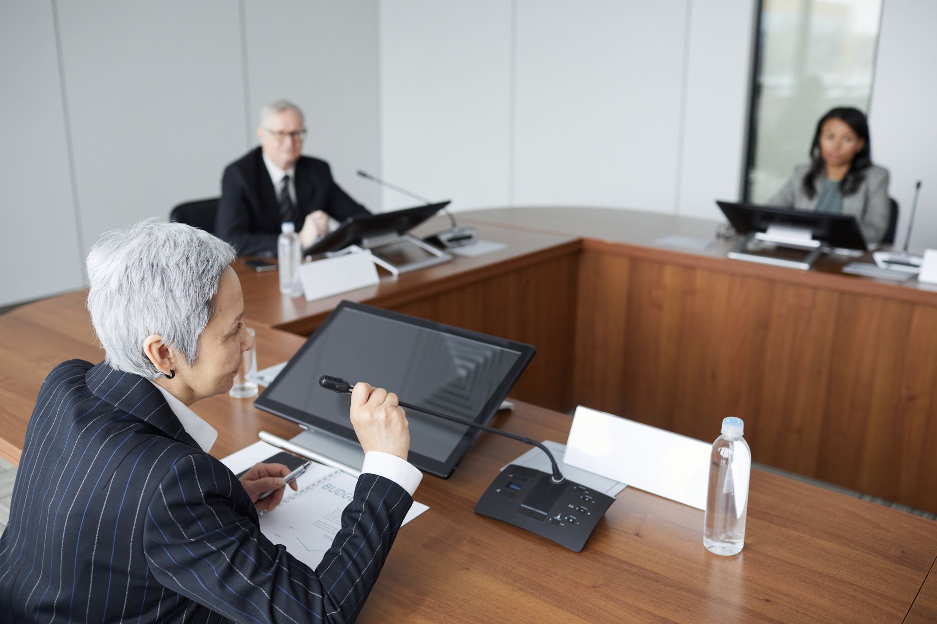 A gray-haired executive in a pinstripe suit speaks into a conference microphone while two colleagues listen across a wooden boardroom table