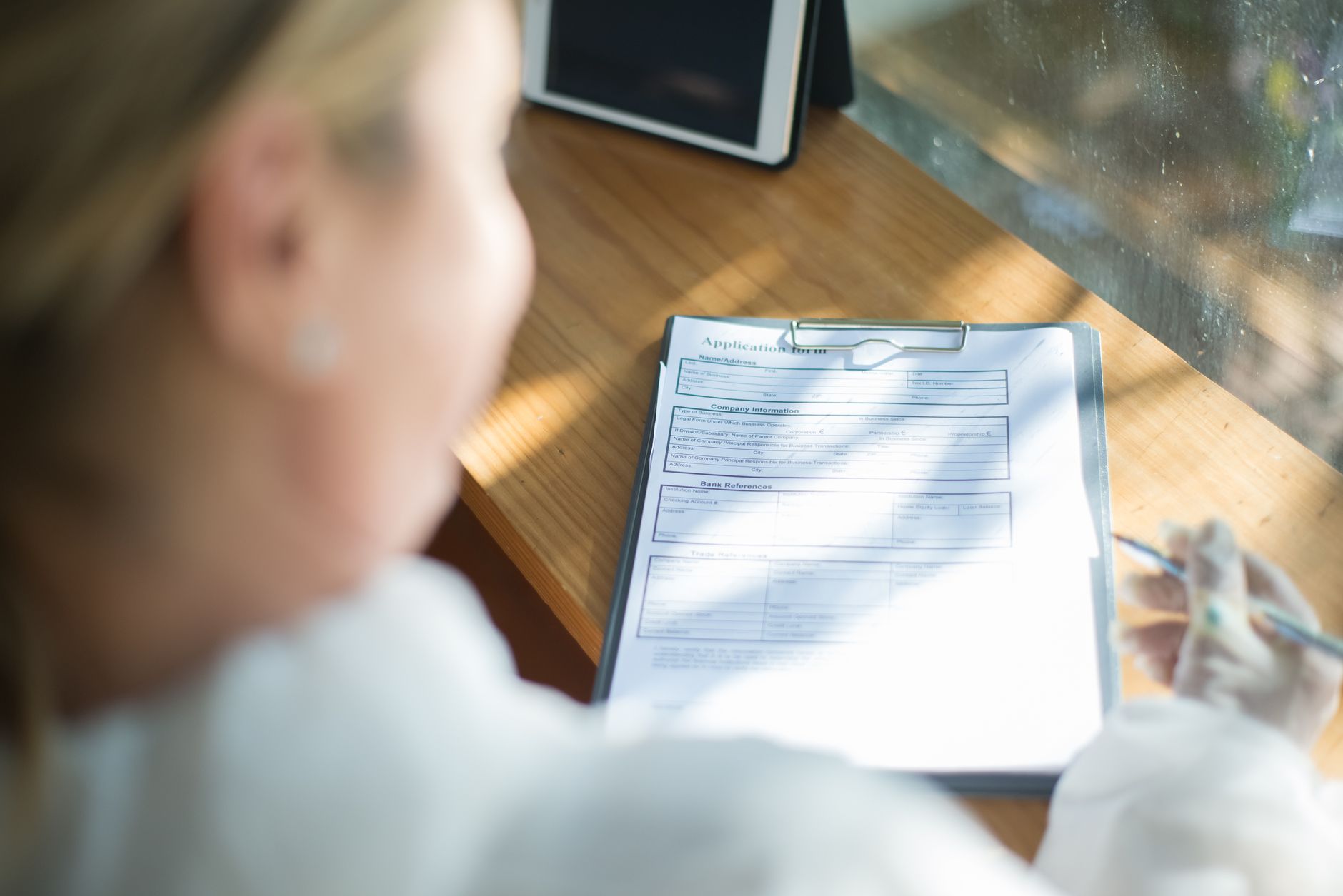 An applicant fills out a paper application form on a clipboard at a sunlit wooden desk