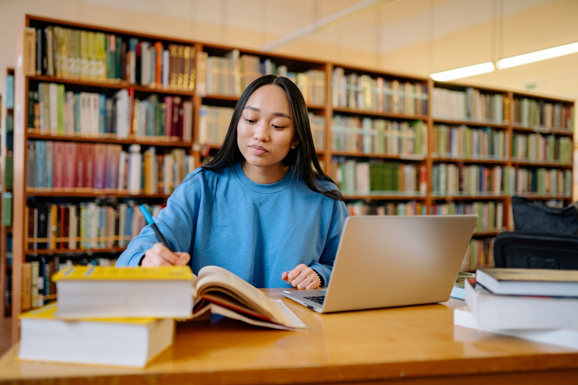 A student in a blue sweatshirt takes notes from a textbook beside an open laptop at a library table lined with bookshelves