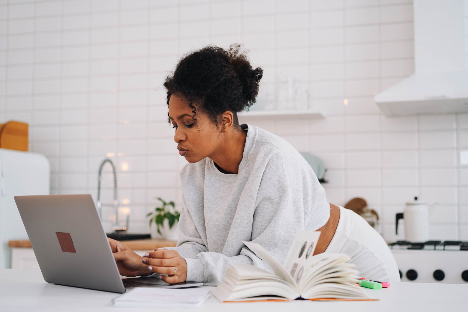 A student works on a laptop at a kitchen counter with an open textbook and highlighters beside her